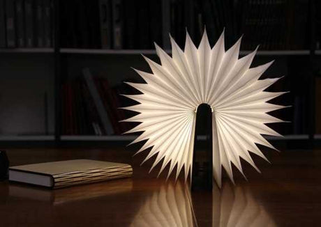 Helles, spitz zulaufendes Lampenobjekt mit sternförmigen Flügeln auf einem Tisch, im Hintergrund Bücherregal.