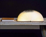 Auf einem weißen Tisch liegen ein braunes Buch und eine aufgeschlagene, beleuchtete Faltlampe.