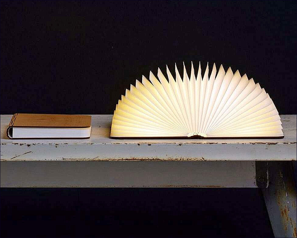 Ein Buch und eine aufgeschlagene Lampe in Form eines Fächers auf einem weißen Tisch vor schwarzem Hintergrund.
