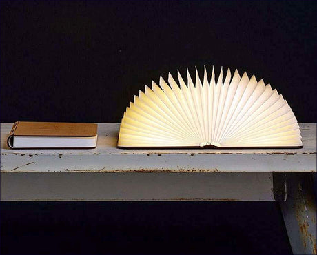 Ein Buch und eine aufgeschlagene Lampe in Form eines Fächers auf einem weißen Tisch vor schwarzem Hintergrund.