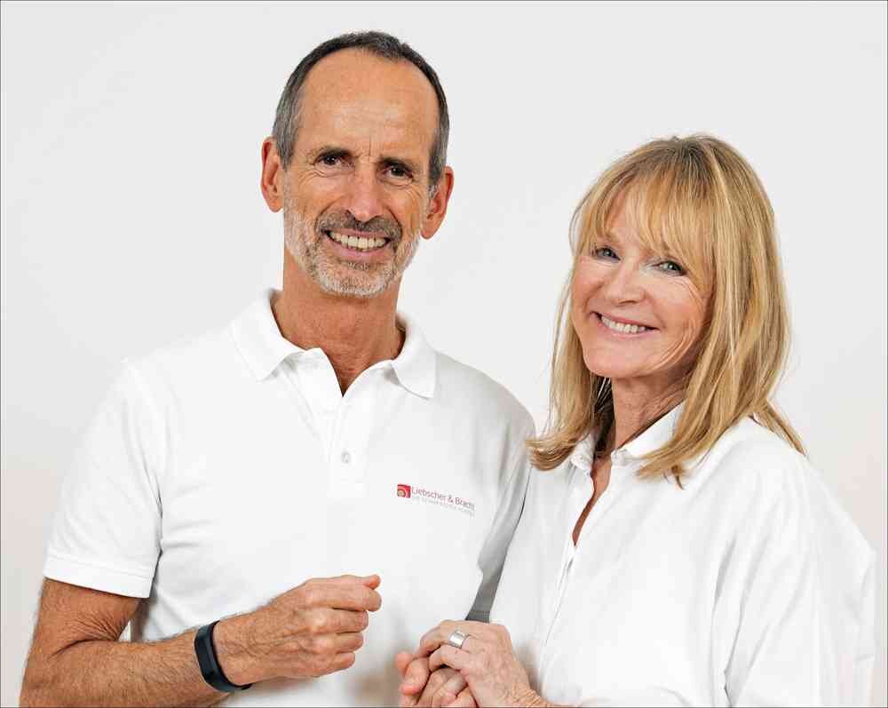 Ein Mann und eine Frau, beide in weißen Hemden, stehen vor weißem Hintergrund und lächeln.