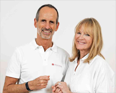 Ein Mann und eine Frau, beide in weißen Hemden, stehen vor weißem Hintergrund und lächeln.