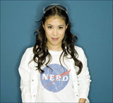 Junge Frau mit dunklen, lockigen Haaren, Brille auf Kopf, trägt weißes T-Shirt mit NASA-Logo und weißen Jacke vor blauer Wand.