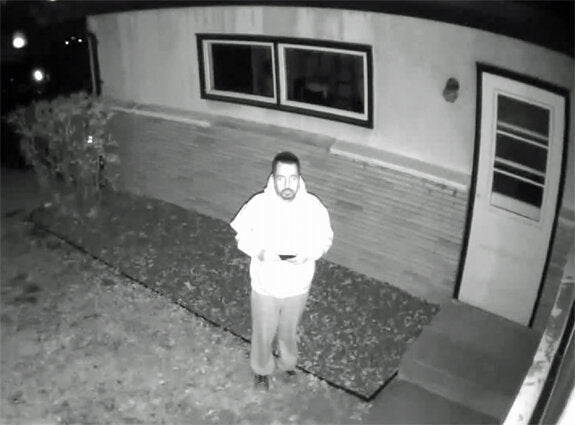 Mann steht vor Haus, hält Smartphone, nachts, schwarz-weiß, vor Fenster und Tür.