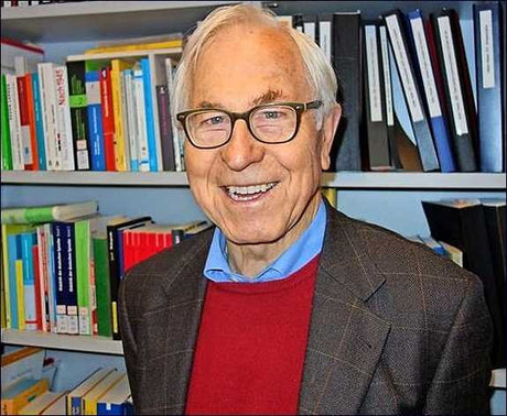 Älterer Mann mit Brille, grauem Haar, lächelnd, in einem Büro mit Bücherregal im Hintergrund.