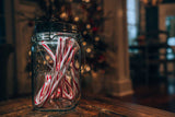 Glas mit Weihnachtslutschern auf einem Holztablett, im Hintergrund Weihnachtsbaum mit Lichtern.