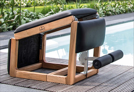 Fitnessgerät mit Holzrahmen, schwarzer Polsterung und Griff, neben Pool und Pflanzen.