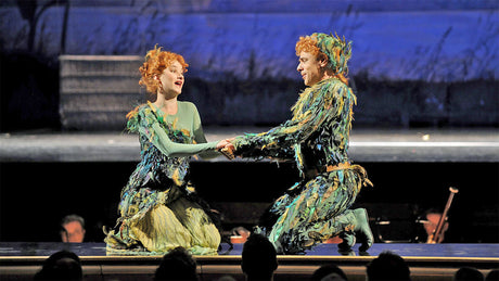 Zwei Schauspieler in bunter, tierähnlicher Kostümierung auf der Bühne, halten Händchen und sprechen.