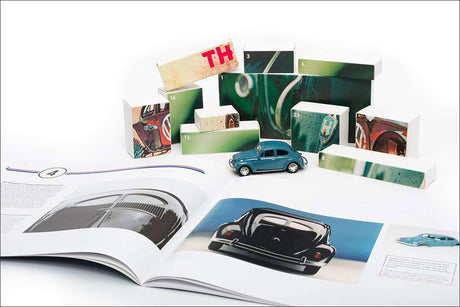 Offener Katalog mit Abbildungen eines schwarzen Autos und eines blauen VW Käfers, umgeben von Modellauto und bunten Bausteinen.