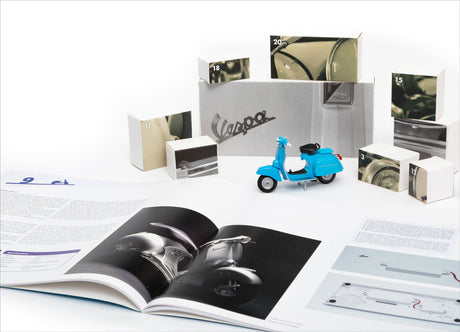 Kleine blaue Vespa, offene Broschüre, Adventskalender mit Vespa-Bildern, und Vespa-Logo auf Pappkarton.