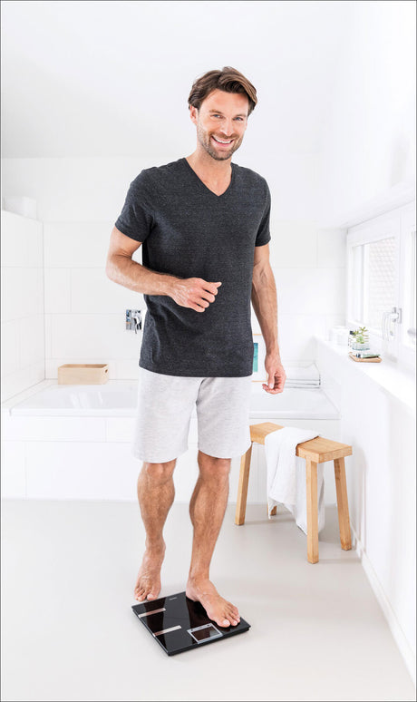 Mann steht auf einer Waage in einem weißen Badezimmer, trägt T-Shirt und Shorts, lächelt.