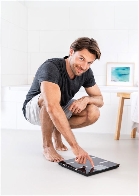 Mann steht auf Waage, zeigt auf Display, in weißem Raum mit Bild an Wand, sitzt auf Zehenspitzen, trägt T-Shirt und Shorts.