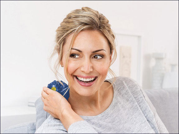 Frau lächelt, hält eine blaue Wärmflasche an die Schulter, sitzt auf einem Sofa in einem hellen Raum.