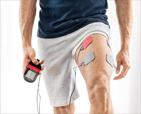 Mann mit EKG-Elektroden an Oberschenkel, hält EKG-Gerät in Hand, trägt graue Shorts und blauen T-Shirt.