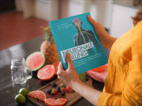Person hält ein Buch mit dem Titel "Neuroale Heilung" vor einer Obstschale mit Wassermelone, Grapefruit, Limetten und Beeren.