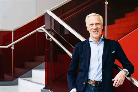 Älterer Mann mit weißem Haar, blauer Hemd, dunklem Blazer, steht an rotem Treppengeländer in moderner Umgebung.