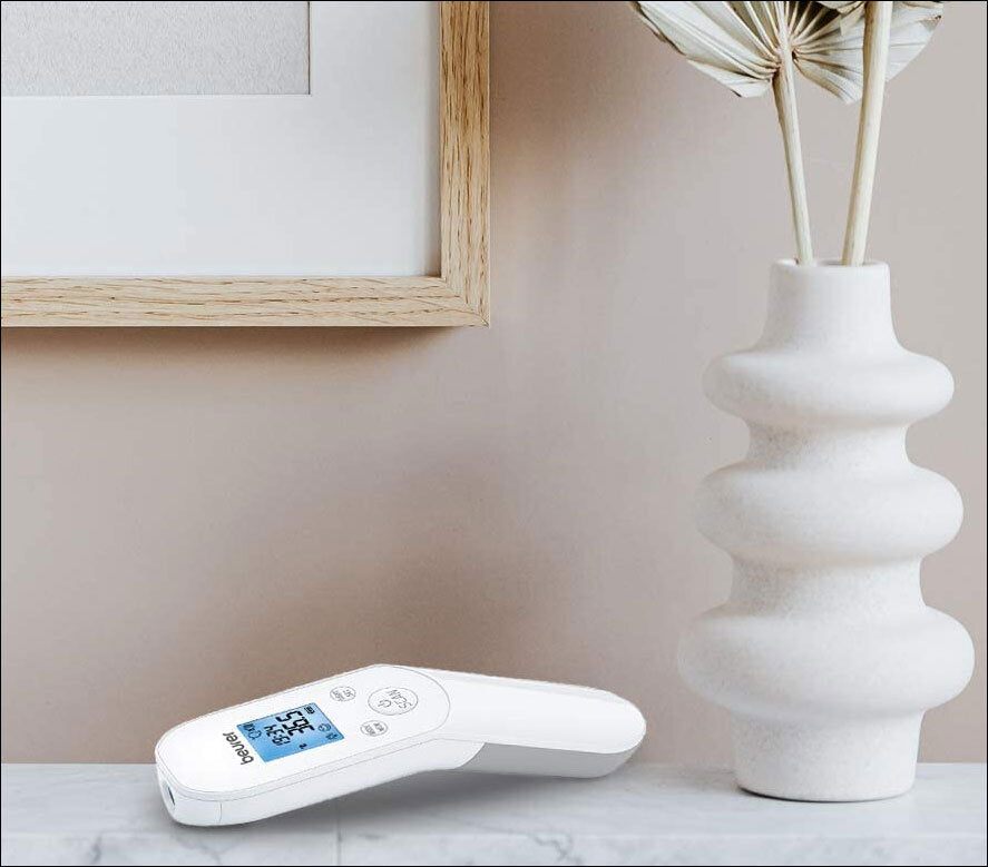 Digitale Fieberthermometer liegt auf einer weißen Oberfläche neben einer weißen Vase mit getrockneten Zweigen.