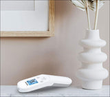 Digitale Fieberthermometer liegt auf einer weißen Oberfläche neben einer weißen Vase mit getrockneten Zweigen.