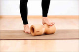 Person steht auf einem Holzroller auf Yoga-Matte, trägt schwarze Leggings, im Hintergrund weiße Wand.