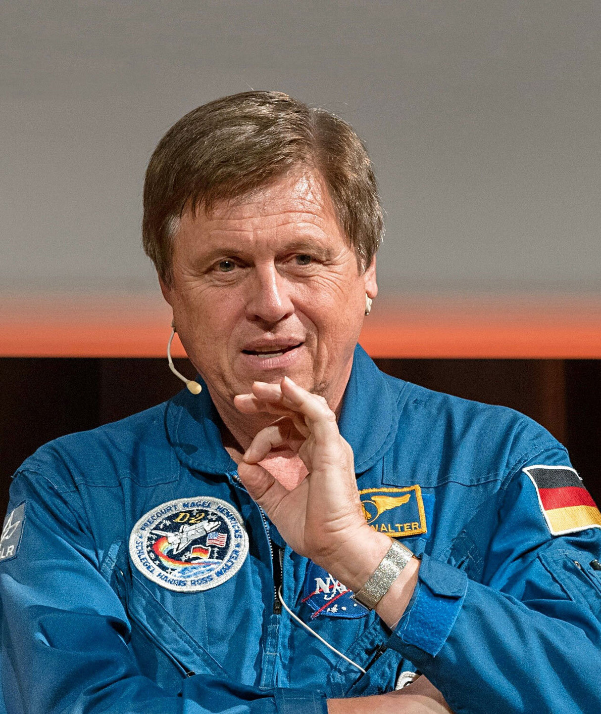 Mann in NASA-Autoanzug mit Abzeichen, spricht mit Headset, gestikuliert mit Hand.