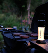 Grill mit vier Patties, im Hintergrund Bäume und Pflanzen, im Vordergrund eine LED-Lampe auf dem Grill.