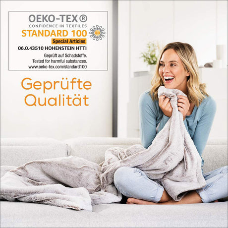 Frau sitzt auf Sofa, hält eine Decke und lacht. Text auf Bild: "Geprüfte Qualität" und Zertifikat "Oeko-Tex Standard 100".