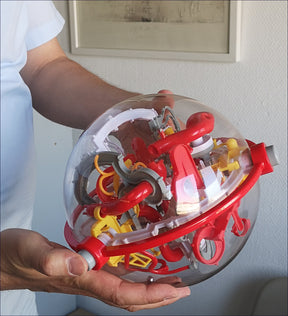 Hand hält eine transparente Kugel mit komplexen roten, gelben und grauen Kunststoffteilen.
