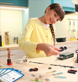 Junge Frau mit brauner Zopf, gelber Pullover, arbeitet an Bastelprojekten auf einem Tisch mit verschiedenen Materialien.