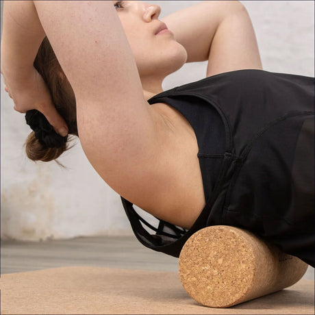 Frau liegt auf einem Yoga-Roller, hebt Arme hinter den Kopf, trägt schwarze Sportkleidung, im Hintergrund weiße Wand.