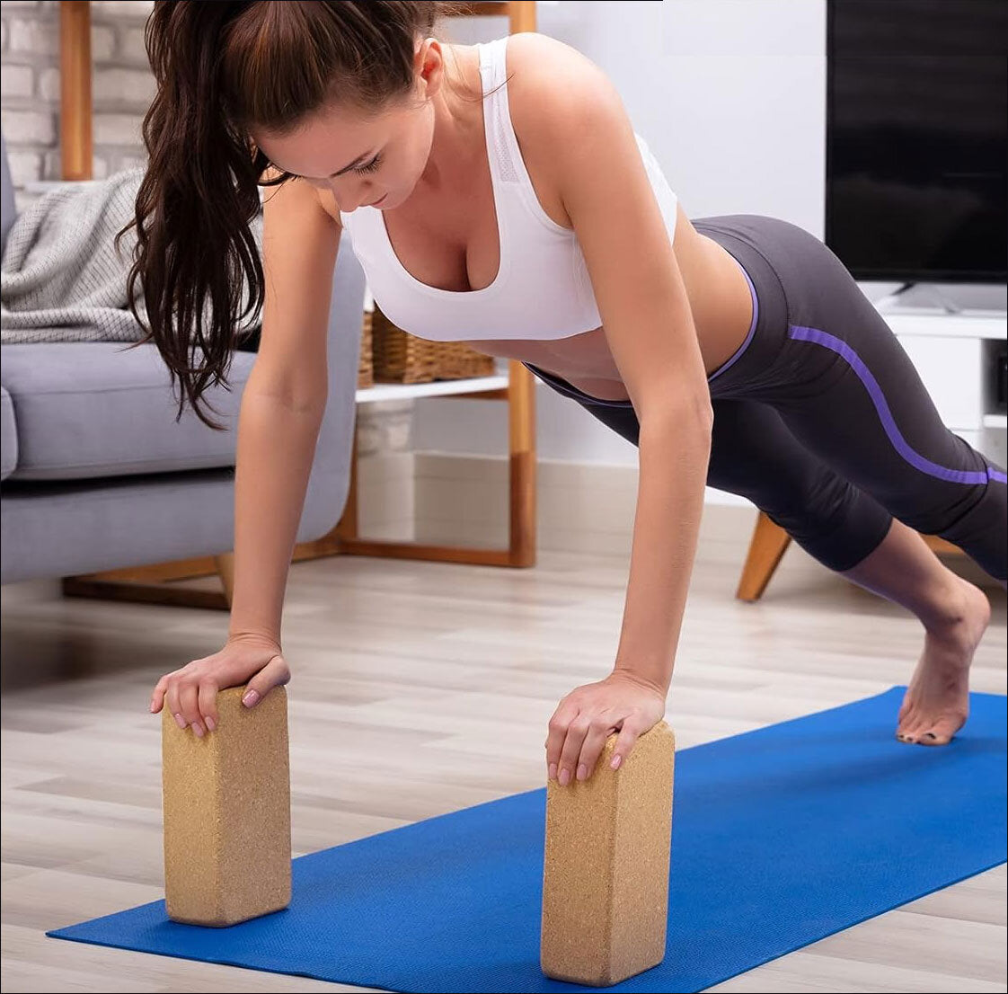 Frau macht Yoga im Wohnzimmer, in Plank-Position mit Yoga-Matte und Ziegelblöcken.