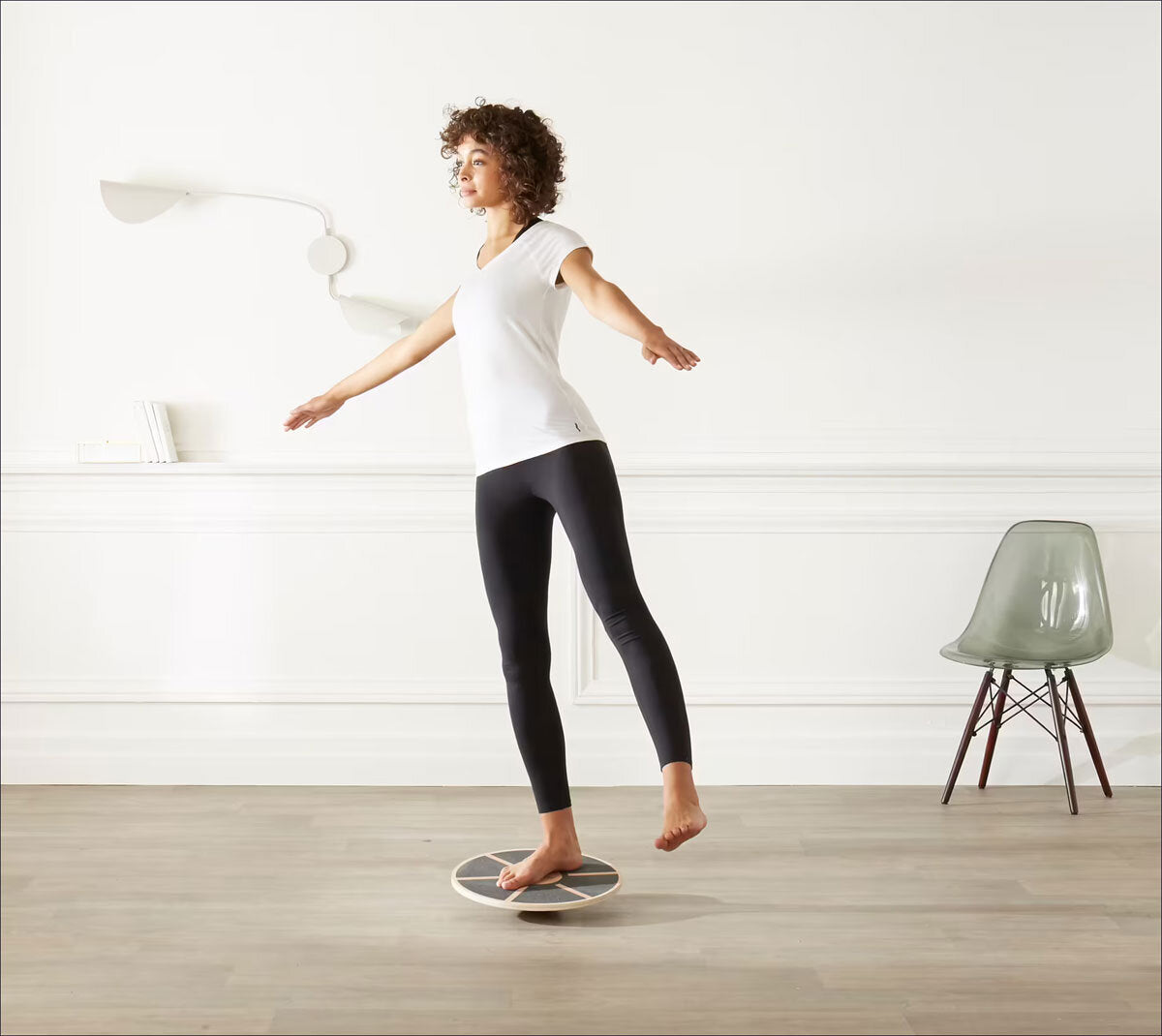 Frau steht auf einem Balance-Board, Arme seitlich ausgebreitet, in einem weißen Raum mit Stuhl und Lampe.