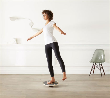 Frau steht auf einem Balance-Board, Arme seitlich ausgebreitet, in einem weißen Raum mit Stuhl und Lampe.