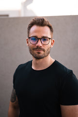 Porträt eines Mannes mit Brille, dunklem T-Shirt, kurzer Haarfrisur und Bart, vor grauem Hintergrund.