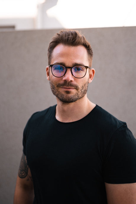 Porträt eines Mannes mit Brille, dunklem T-Shirt, kurzer Haarfrisur und Bart, vor grauem Hintergrund.