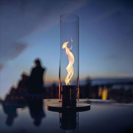 Brennender Kerzenflamme in einem hohen Glas, im Hintergrund unscharfer Mensch bei Sonnenuntergang.
