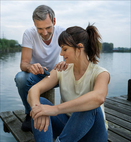 Frau wird am Arm von einem Mann mit einem Dermatoskop untersucht, während sie auf einem Steg am Wasser sitzt.