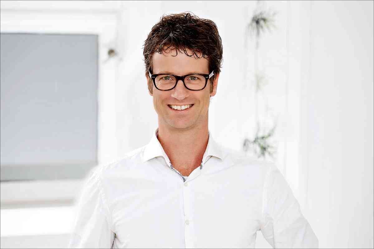 Mann mit dunklen, lockigen Haaren, Brille und weißem Hemd, lächelnd vor weißem Hintergrund.