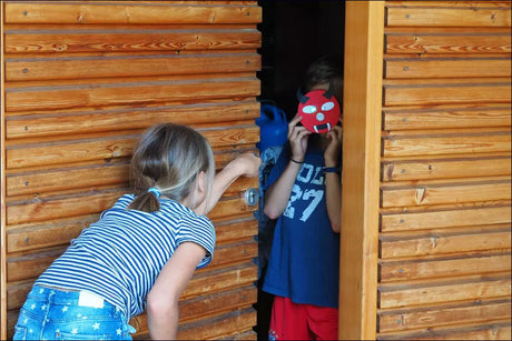 Zwei Kinder hinter einer Holztür, eines mit einem roten Dämonen-Masken, das andere reicht durch den Türspalt.
