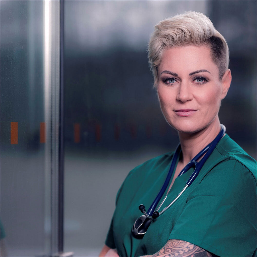 Ärztin in grüner Uniform mit Stethoskop, kurze blonde Haare, Tattoo am Arm, vor einem medizinischen Hintergrund.