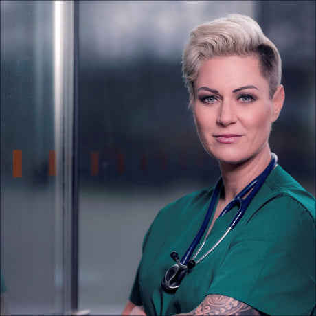 Ärztin in grüner Uniform mit Stethoskop, kurze blonde Haare, Tattoo am Arm, vor einem medizinischen Hintergrund.