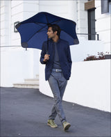 Mann mit dunklem Haar, blauer Jacke, grauem Pullover, grauen Hosen, grünen Schuhen, hält blauen Regenschirm