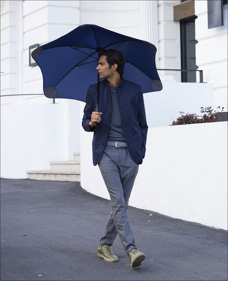 Mann mit dunklem Haar, blauer Jacke, grauem Pullover, grauen Hosen, grünen Schuhen, hält blauen Regenschirm