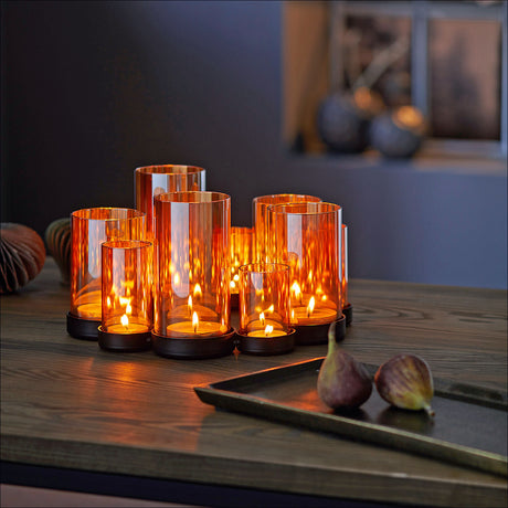 Mehrere orange Kerzen in Gläsern auf einem Holztisch, daneben eine Schale mit zwei Feigen.