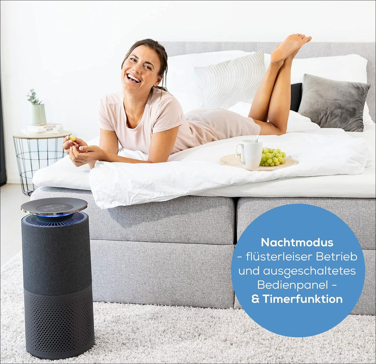 Frau liegt lachend auf einem Bett, neben einem Luftreiniger mit Display, auf dem eine blaue Anzeige leuchtet.