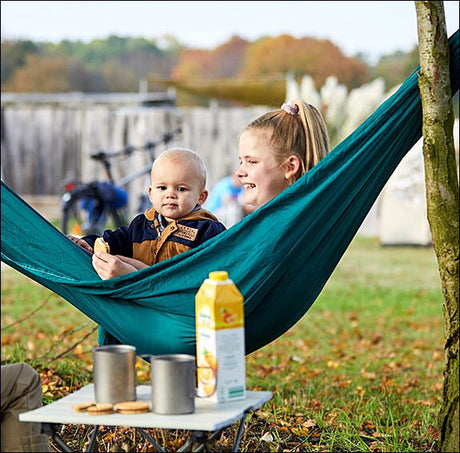 Eine Frau und ein Baby sitzen in einer Hängematte im Garten, im Vordergrund ein Tisch mit Getränken und Keksen.
