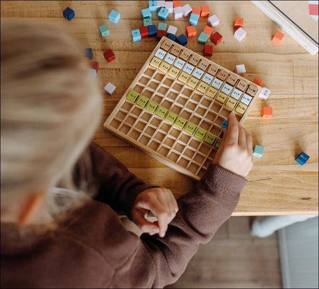 Kind eines Kindes spielt mit einem Holz-Excel-ähnlichen Spielbrett und bunten Würfeln auf einem Tisch.