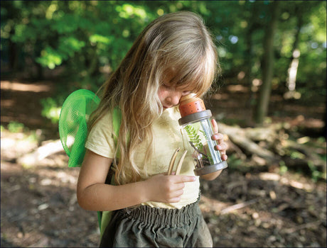 Mädchen mit Rucksack und Lupe im Wald, betrachtet eine Pflanze in einem transparenten Behälter.