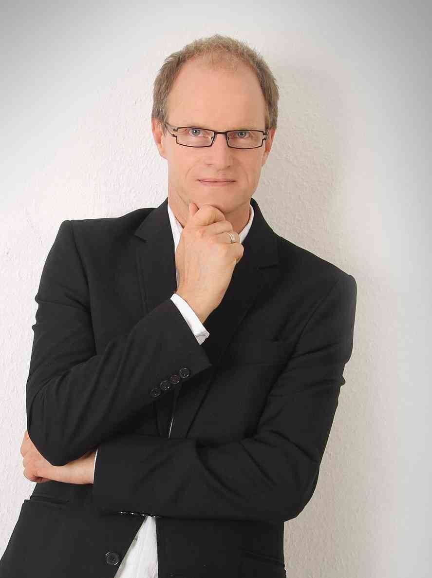 Mann mit Brille, schwarzem Anzug und weißem Hemd, nachdenklich mit Hand am Kinn vor weißer Wand.