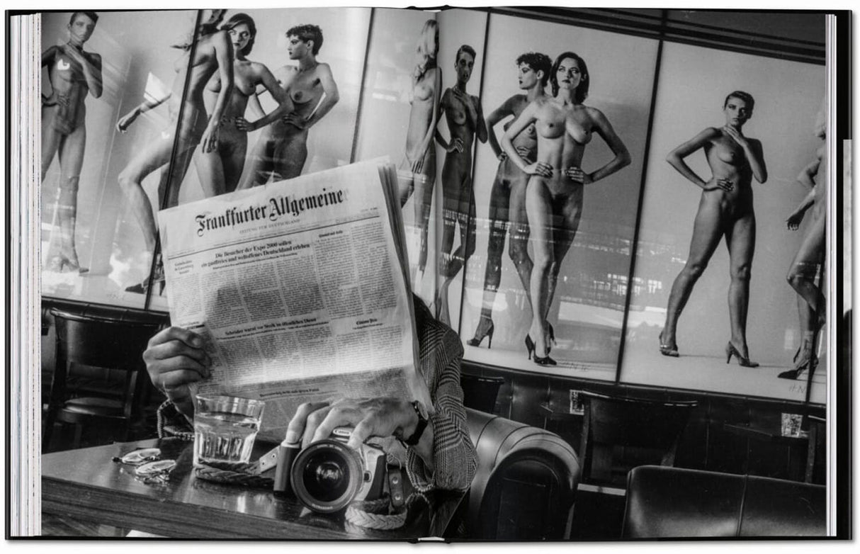 Person liest Zeitung vor Spiegel mit Akt-Fotografien, Kamera und Glas Wasser auf Tisch.