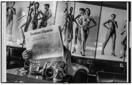 Person liest Zeitung vor Spiegel mit Akt-Fotografien, Kamera und Glas Wasser auf Tisch.